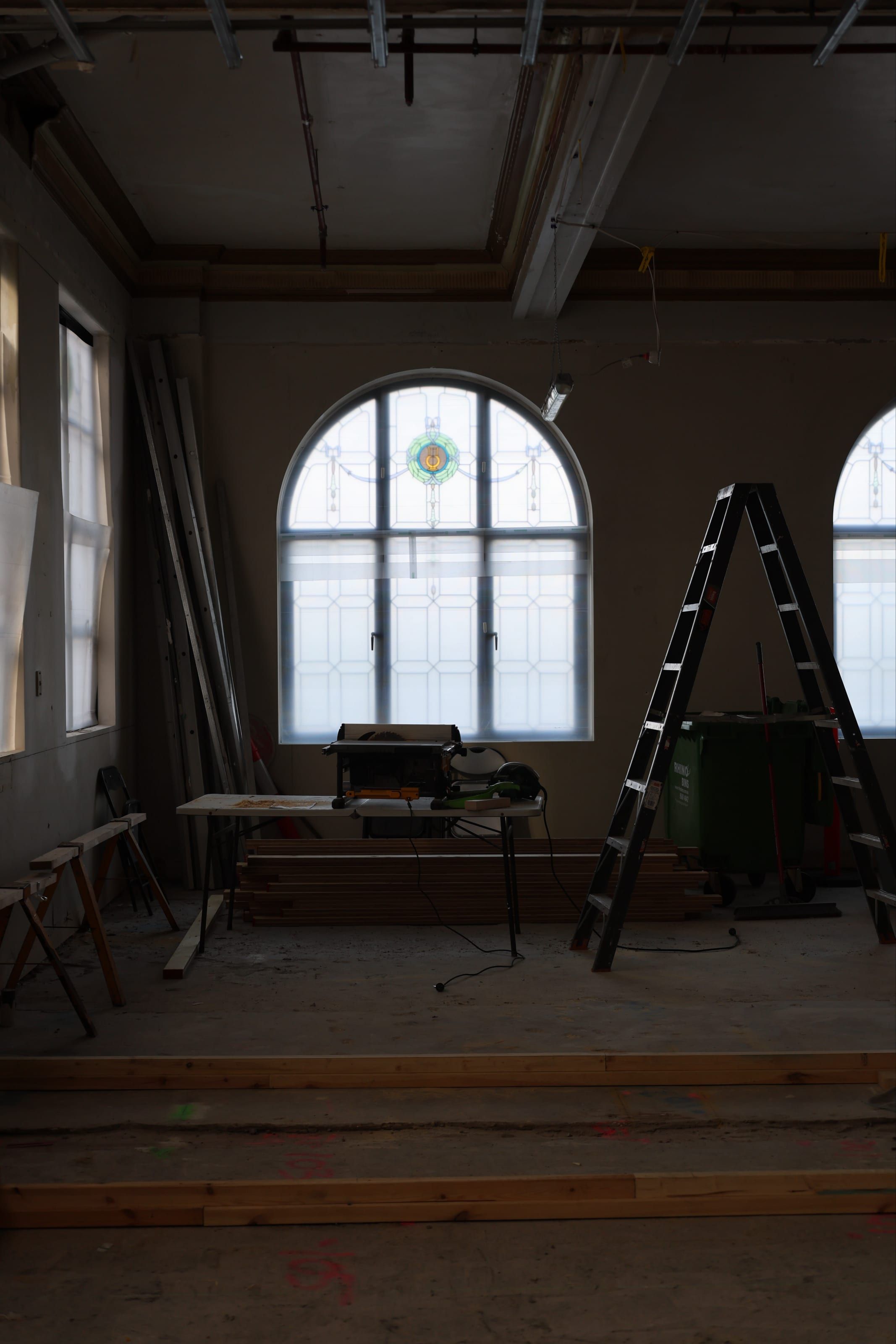 An arched leadlight window with a central crest, framed by stacked timber and a drop saw